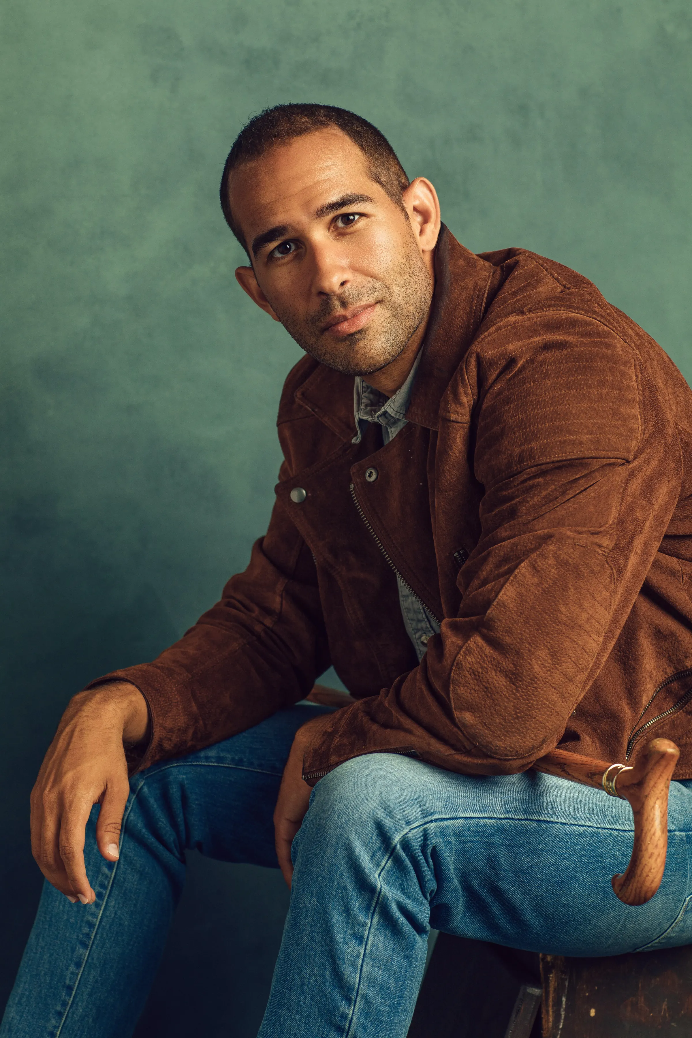 Portrait of Andrew Reid in a brown suede jacket, seated against a teal backdrop with his cane.